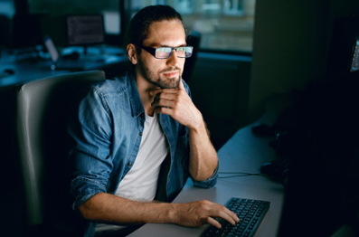 Male with glasses sitting in a computer with two monitors in a dark workspace